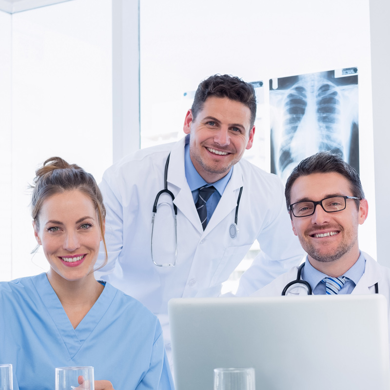 Three smiling healthcare professionals, including two male doctors in lab coats and a female medical professional in blue scrubs, pose around a laptop in a clinical setting with X-ray images visible behind them.